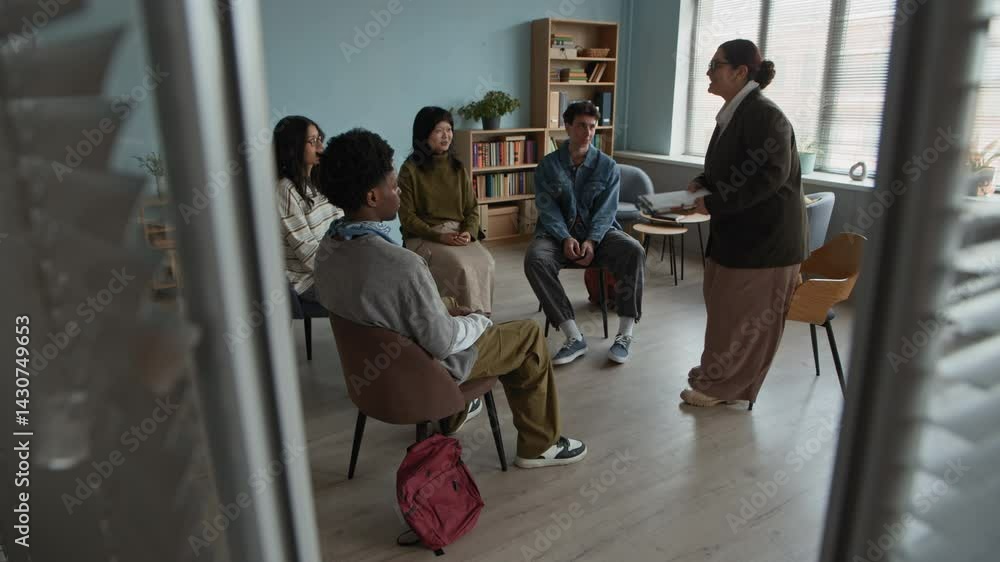 Full shot of diverse group of students sitting in circle in cozy classroom with young biracial female psychologist entering room, taking seat and introducing herself starting group therapy Full shot of diverse group of students sitting in circle in cozy classroom with young biracial female psychologist entering room, taking seat and introducing herself starting group therapy
