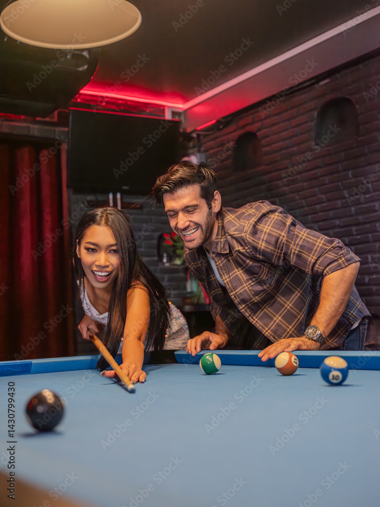 Two young adults gathered around a pool table in a modern bar setting. A woman lines up her shot with focused concentration while a male friend steadies the cue ball for her.
