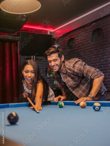 Obraz na płótnie Two young adults gathered around a pool table in a modern bar setting. A woman lines up her shot with focused concentration while a male friend steadies the cue ball for her. w salonie