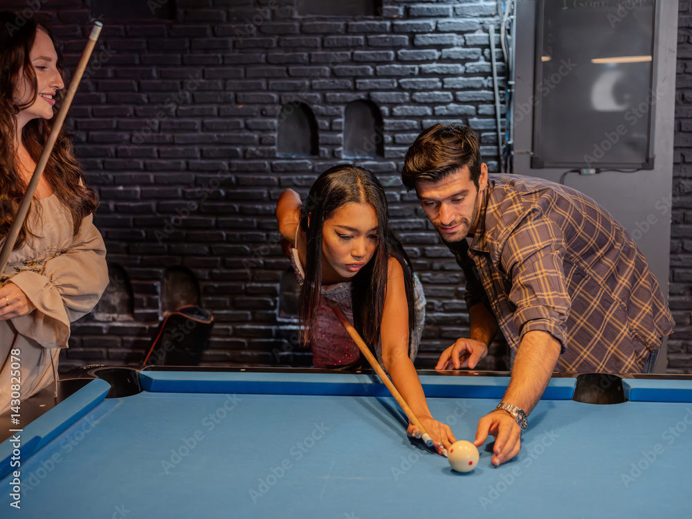 Three young adults gathered around a pool table in a modern bar setting. A woman lines up her shot with focused concentration while a male friend steadies the cue ball for her.