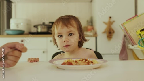 Mother feeding happy child pasta in bright kitchen at table. Joyful child happily enjoying delicious food, emphasizing importance of family bonding during mealtime
