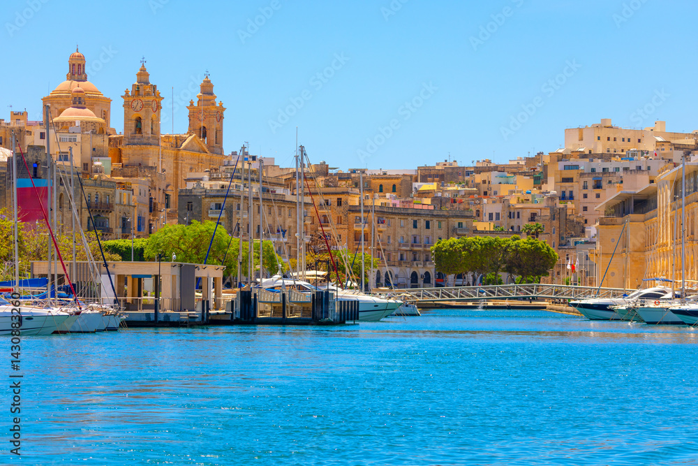 Birgu fortified city on the south side of the Grand Harbour of Malta. Vibrant coastal cityscape features a waterfront with numerous boats and yachts docked along harbor, framed by clear blue water