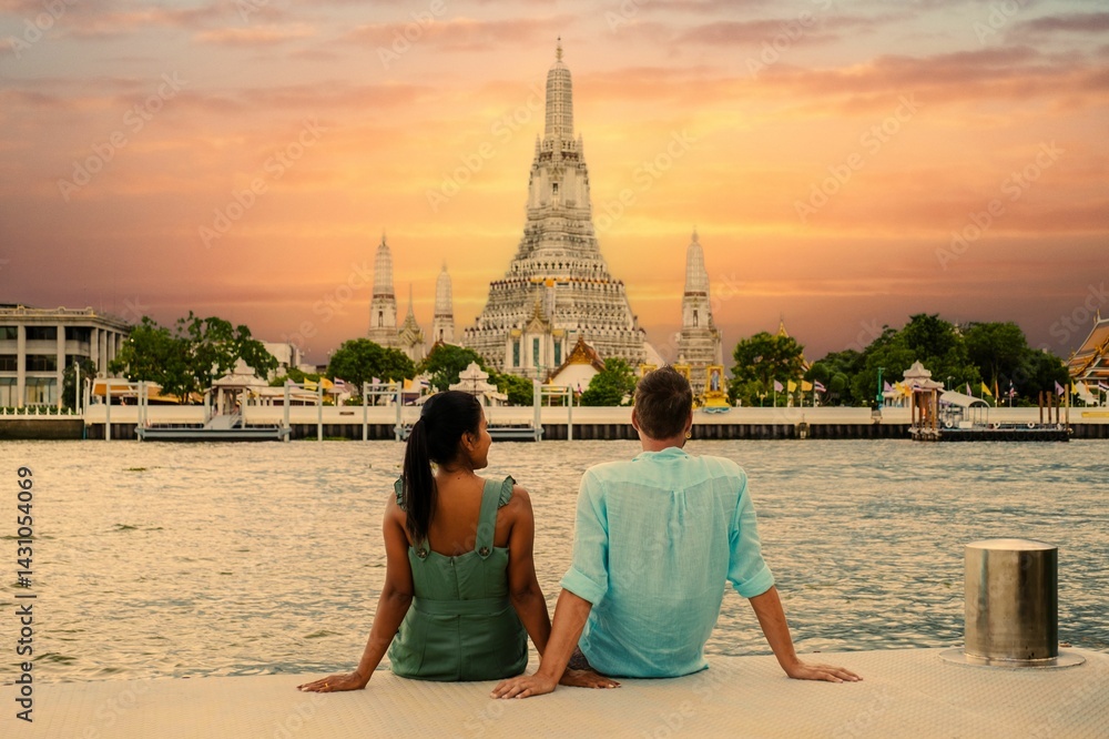 Fototapeta premium A couple relaxes by the Chao Praya River as they admire the stunning sunset view of Wat Arun Temple in Bangkok, Thailand. The vibrant colors of the sky create a romantic atmosphere.