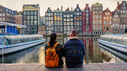 Foto Two people enjoy a serene moment by the canal in Amsterdam, surrounded by charming colorful buildings and gentle boats