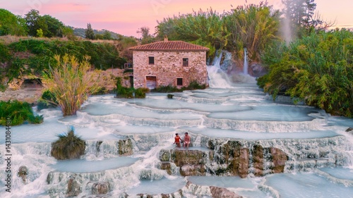 Fényképezés A couple relaxes in Saturnia thermal waters in Italy, surrounded by lush greenery and charming stone buildings as the sun sets, casting golden hues over the beautiful Tuscan landscape