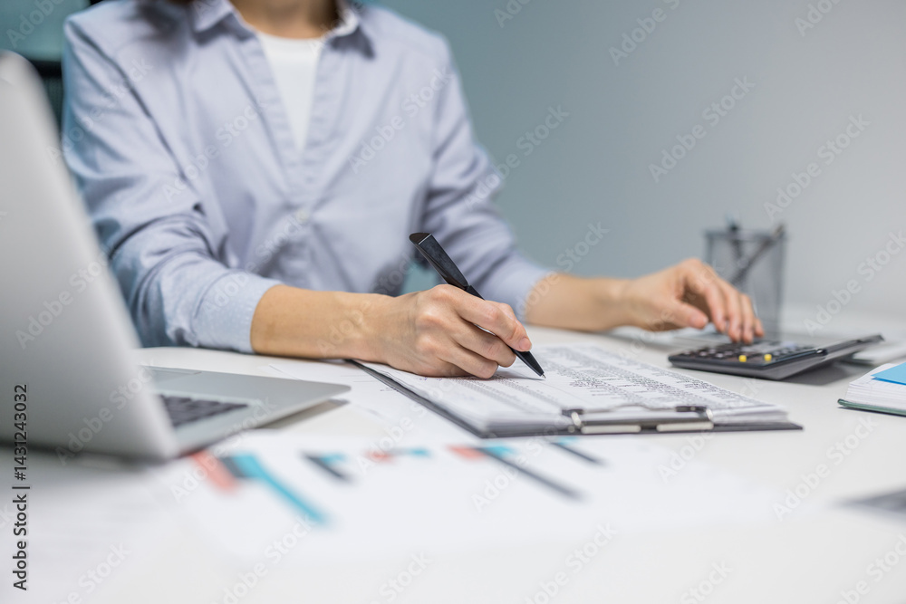 A person diligently works on financial documents at a desk, using a calculator and laptop. The scene focuses on work and accounting activities A person diligently works on financial documents at a desk, using a calculator and laptop. The scene focuses on work and accounting activities