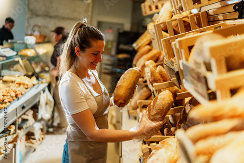 Canvas Print Cheerful saleswoman wearing gloves is arranging loaves of freshly baked bread on