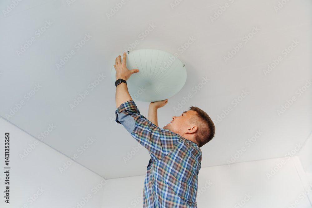 A man in a plaid shirt holds a large white round ceiling lamp in his hands.