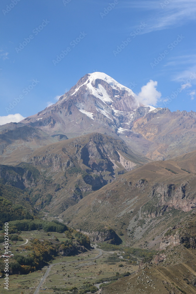 Fototapeta premium Mount Kazbek, a majestic snow-capped peak in Kazbegi, Georgia, towers above the Caucasus mountain landscape. A stunning scene perfect for travel, adventure, nature, and cultural destination themes.