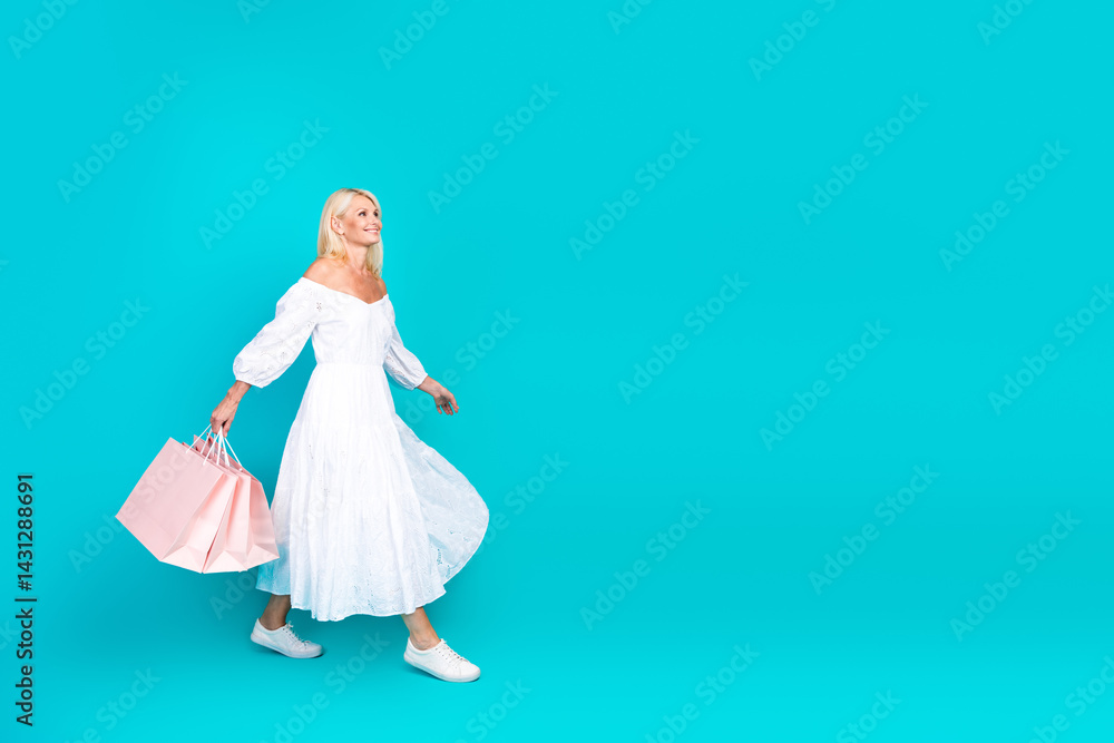 Naklejka premium Mature stylish woman in a white summer dress walking and carrying shopping bags against a vibrant turquoise background