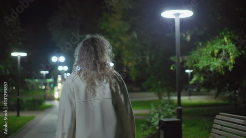 Rear view lady in white coat walking in open park at night, people faintly visible, overhead lamps illuminate surroundings, curly hair gently lit, capturing calm city ambiance with foliage