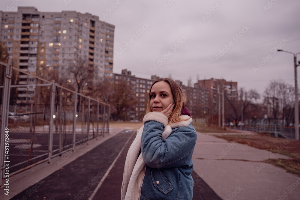 Fototapeta premium Young woman wearing a light jacket stands outside a sports court during twilight in a residential area