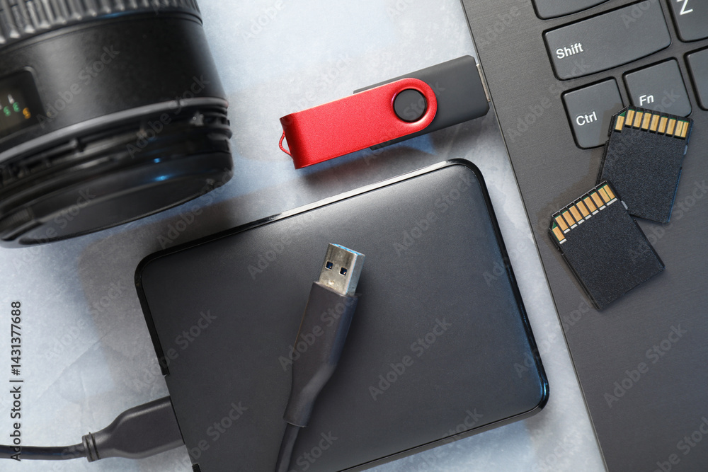 Obraz premium Flash drive connected to laptop, SD memory cards, external hard disk and camera lens on gray textured table, flat lay