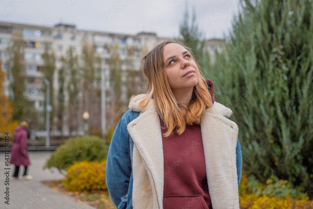 Young woman in a casual outfit stands confidently in an urban park during autumn with colorful trees and modern buildings in the background Young woman in a casual outfit stands confidently in an urban park during autumn with colorful trees and modern buildings in the background