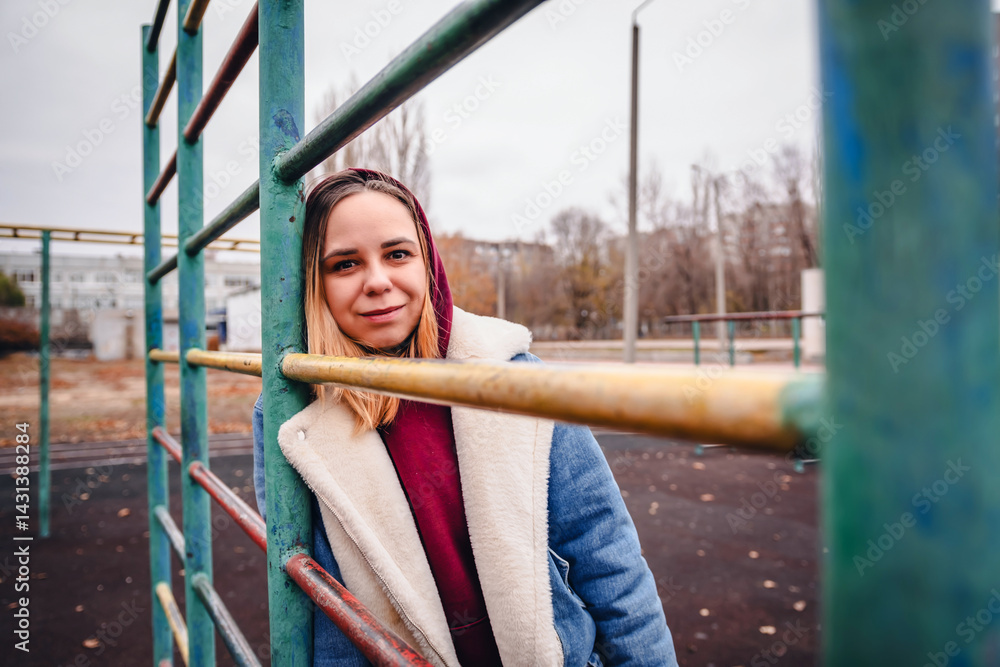 Obraz premium Young woman in a hoodie and coat poses at a playground during autumn in a city park, surrounded by colorful trees and apartment buildings