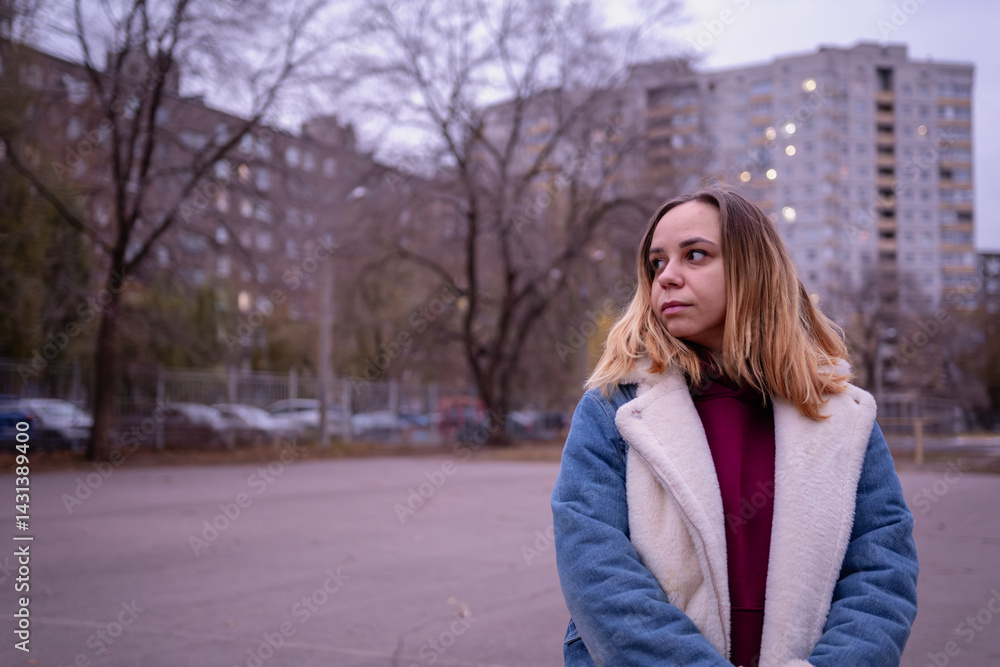 Fototapeta premium Young woman in casual attire stands in an urban park during early evening, surrounded by tall buildings and autumn trees