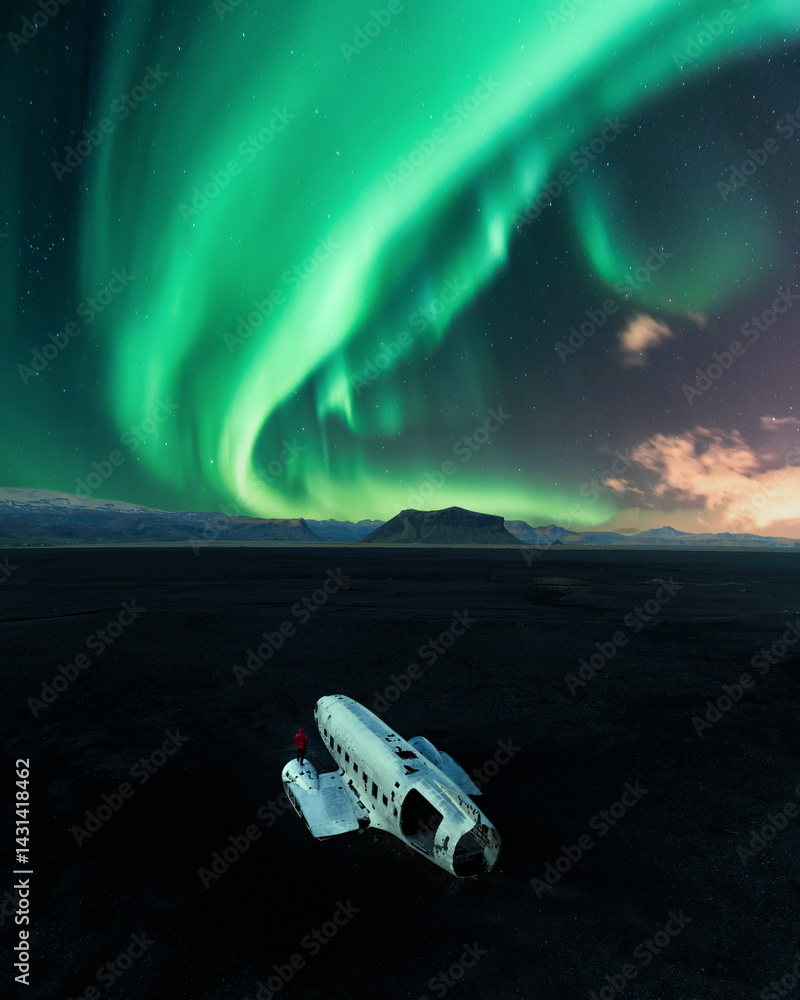Abandoned DC-3 plane wreck on Solheimasandur beach in Iceland beneath vibrant green Northern Lights, with a person in red for scale and contrast.
