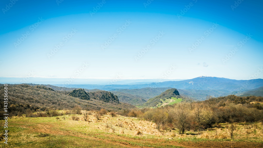 Naklejka premium Serene spring landscape in Kakheti, Georgia, featuring forested hills, lush green meadows, and distant snow-capped peaks under a clear blue sky