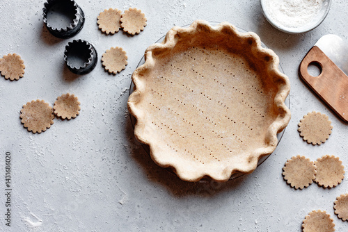 An overhead view of a pie crust and cookie cutouts on a light gray surface, with flour and a dough cutter nearby.