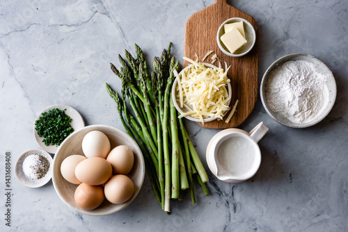 An assortment of cooking ingredients including eggs, asparagus, shredded cheese, butter, flour, milk, chives, and salt and pepper are arranged on a marble countertop.