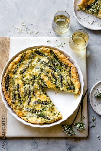 An asparagus and cheese quiche with a slice missing, served alongside glasses of white wine on a marble surface.