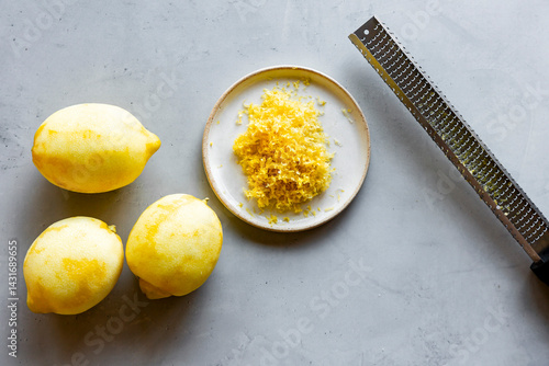 Three lemons, lemon zest on a plate, and a zester on a gray surface.