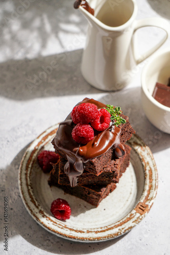 chocolate brownies topped with glossy melted chocolate and fresh raspberries, served on a rustic ceramic plate. The dessert is placed on a textured light grey surface with a white ceramic milk jug