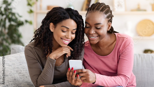 Foto Smiling african american girlfriends checking photos on mobile phone, spending time together at home, copy space
