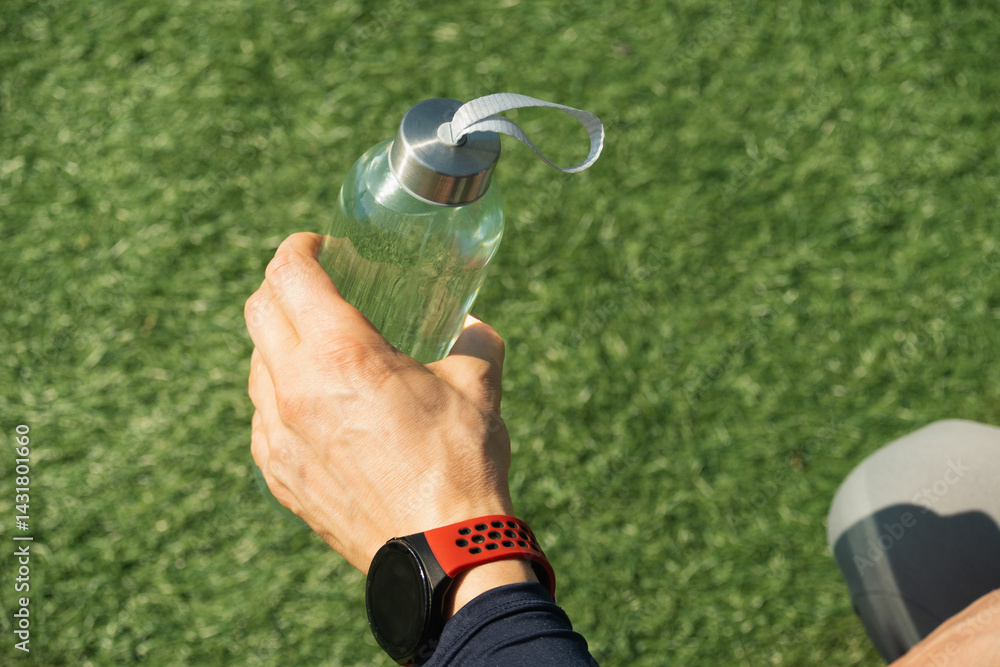 Sport man holding water bottle on outdoor sports ground with green grass. Concept water balance for heat season for workout. Close up. Copy space.