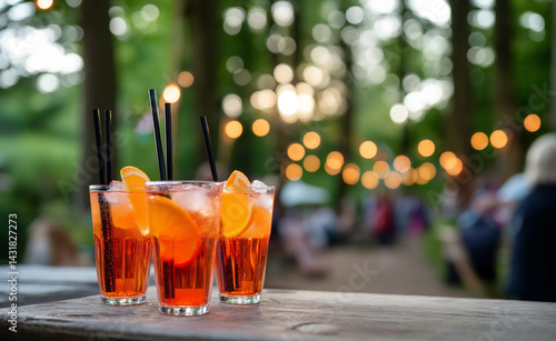 Three refreshing orange cocktails with straws and orange slices on a wooden table at an outdoor summer gathering.
