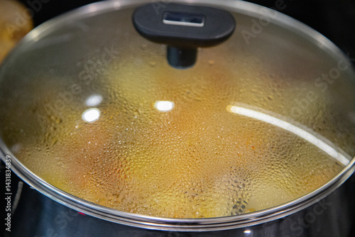 Close-up of a steaming pot with a glass lid during home cooking, showing water condensation and heat reflections, taken in a modern kitchen while preparing a meal.