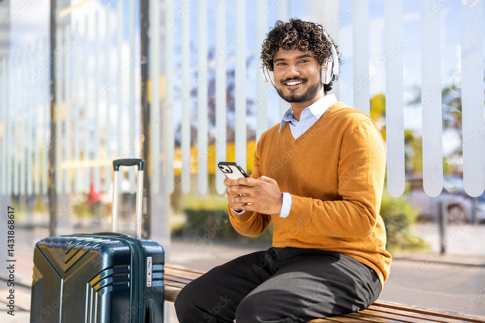 A smiling man with headphones uses his phone while waiting at a bus stop, a suitcase beside him. He is dressed casually and looks relaxed
