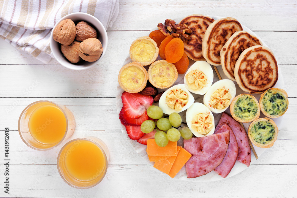 Breakfast or brunch grazing board. Collection of items including fruit, ham, quiche, pancakes and eggs. Top view table scene on a white wood background.