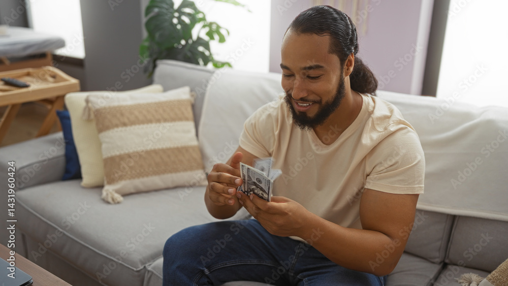 Young man with beard counting money sitting in cozy living room with sofa and pillows around offering a relaxed home atmosphere.