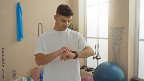 Young man checking smartwatch in a gym wearing casual white shirt, surrounded by fitness equipment, illustrating a modern healthy lifestyle in an indoor sports facility setting.