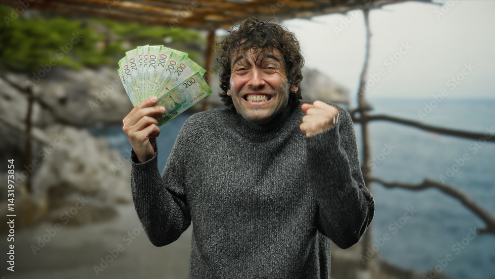 Hispanic man joyfully holding russian roubles on a seaside beach, expressing excitement outdoors in a sweater.