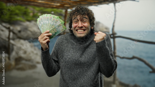Obraz na płótnie Hispanic man joyfully holding russian roubles on a seaside beach, expressing excitement outdoors in a sweater. w salonie
