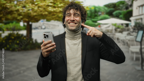 Young hispanic man smiling while pointing at his teeth with a finger and holding a phone in a street setting outdoors, exuding confidence and style in a city environment.