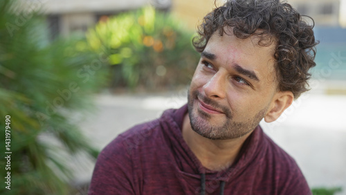 Handsome man with curly hair smiling in an urban park, surrounded by greenery, capturing a serene moment in a bustling city environment, sunlight gently illuminating his face.