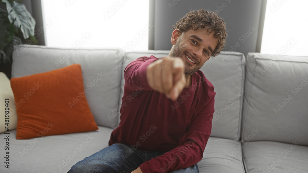 Young man sitting on a couch in a cozy living room pointing at the camera, conveying a friendly and inviting atmosphere in a modern apartment setting. Young man sitting on a couch in a cozy living room pointing at the camera, conveying a friendly and inviting atmosphere in a modern apartment setting.