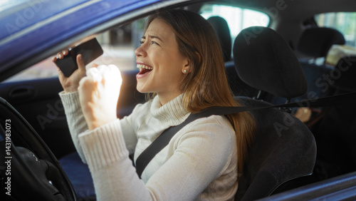 Young woman sitting in a car on an urban street outdoors, smiling and celebrating with a phone in hand, showcasing joy and modern lifestyle in the city.