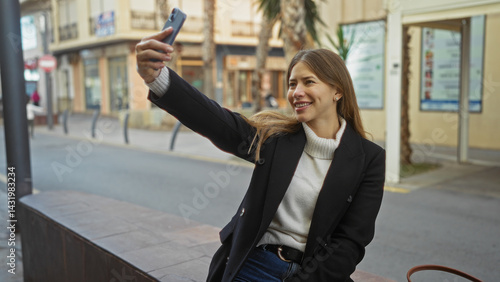 Obraz na płótnie Woman taking selfie on urban street with phone in hand, outdoor setting, wearing stylish jacket, smiling attractively, enjoying cityscape around her. w salonie