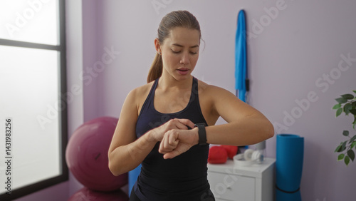 Obraz na płótnie Woman checking smartwatch in gym with focus on time and fitness surrounded by exercise equipment and natural light from window on a bright day indoors. w salonie