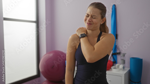 Obraz na płótnie Woman experiencing shoulder pain in a gym, wearing athletic wear, surrounded by fitness equipment, conveying discomfort and effort in a sports center environment. w salonie