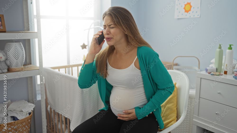 Pregnant woman sitting in a bedroom on phone wearing casual clothing beside baby cradle expressing concern indoors sunlight streaming tattooed hands holding stomach