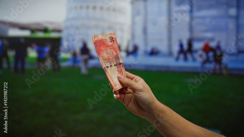 Obraz na płótnie Man holding south african rand banknote outdoors with pisa's iconic architecture in the background, blending global finance and tourism in an urban cityscape. w salonie