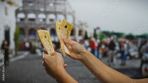 Obraz na płótnie Hands holding philippine pesos in front of the roman coliseum, illustrating investment and travel themes in an urban outdoor setting. w salonie