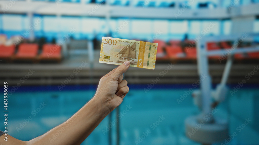 Man holding norwegian krone banknote near outdoor pool at resort, showcasing currency in a leisure setting with blue water backdrop and bright vacation atmosphere.