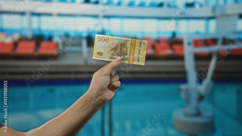 Man holding norwegian krone banknote near outdoor pool at resort, showcasing currency in a leisure setting with blue water backdrop and bright vacation atmosphere.