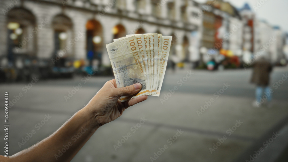 Hand holding danish krone banknotes outdoors in a city street setting, highlighting financial exchange against urban backdrop, with man's figure and historic architecture visible.